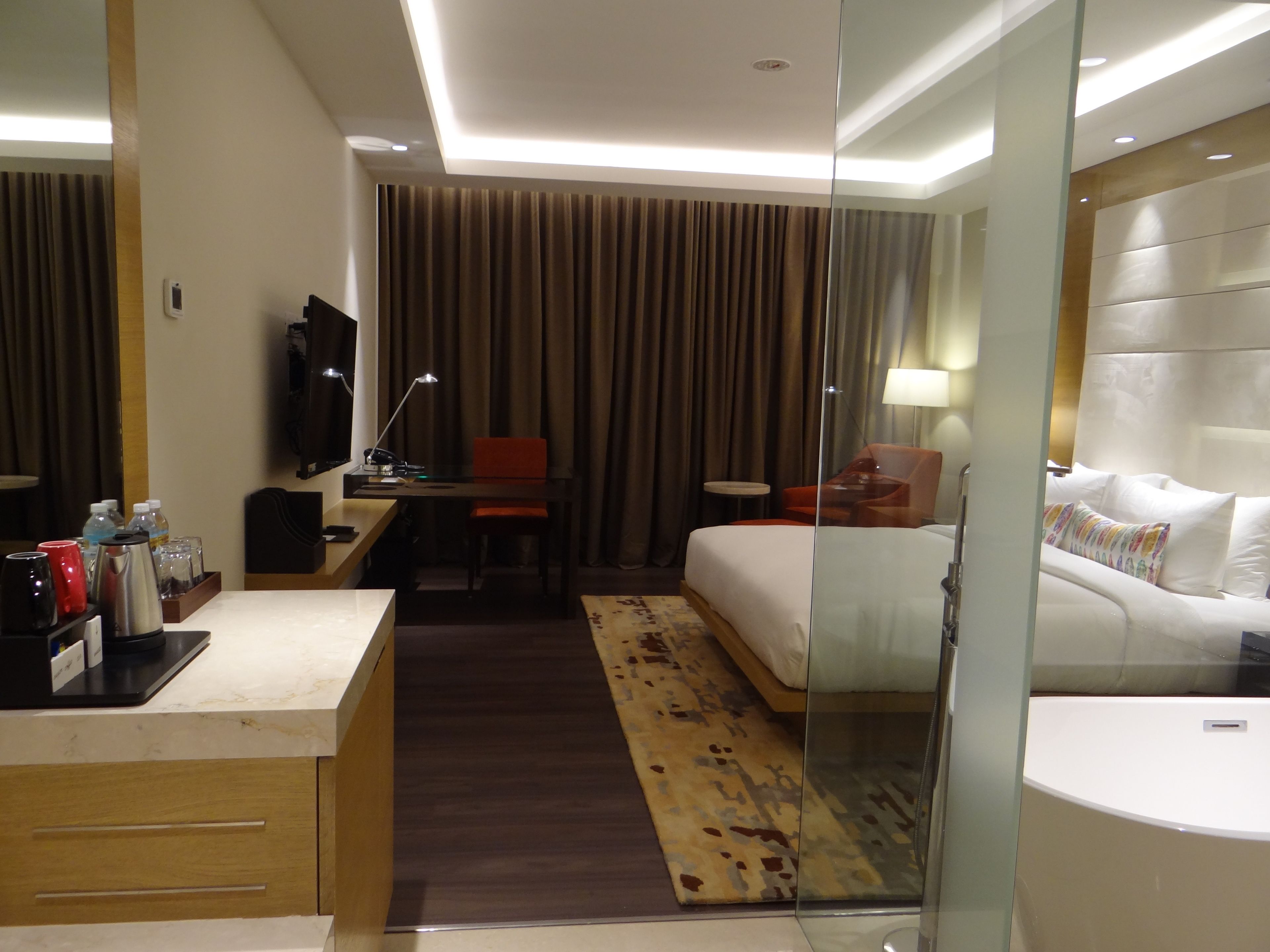 Quarto de hotel com cama, escrivaninha, cadeira, televisão e divisória de vidro.