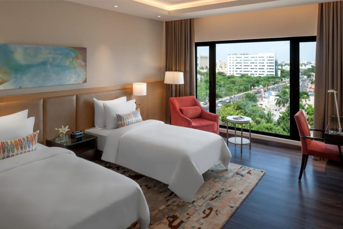 Een hotelkamer met twee bedden, een rode stoel, een klein tafeltje en een groot raam met uitzicht over de stad.