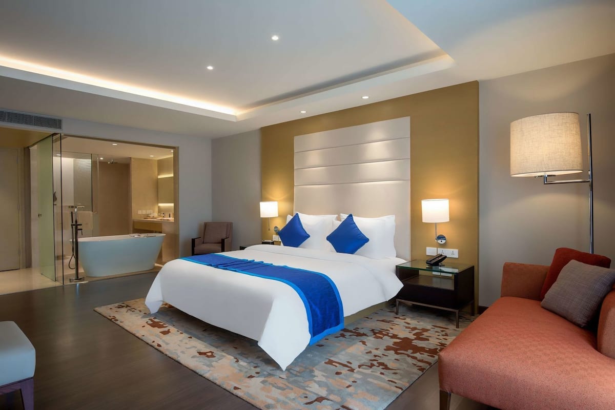 A modern hotel room with a large bed, bedside tables, a nightstand, a lamp, a chair, a glass shower enclosure, and a carpeted floor.
