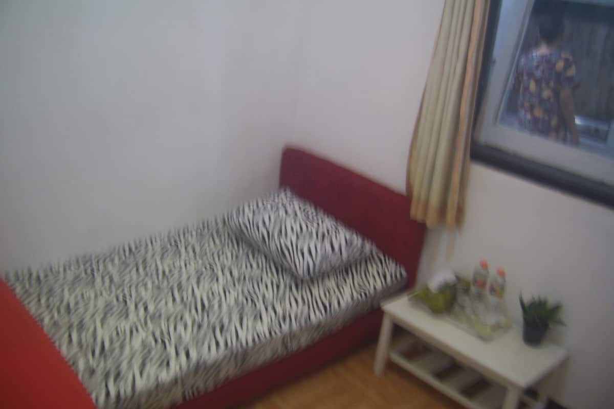 A small, red sofa with a patterned cover, a white side table with a plant and a bottle, and a framed picture on the wall.