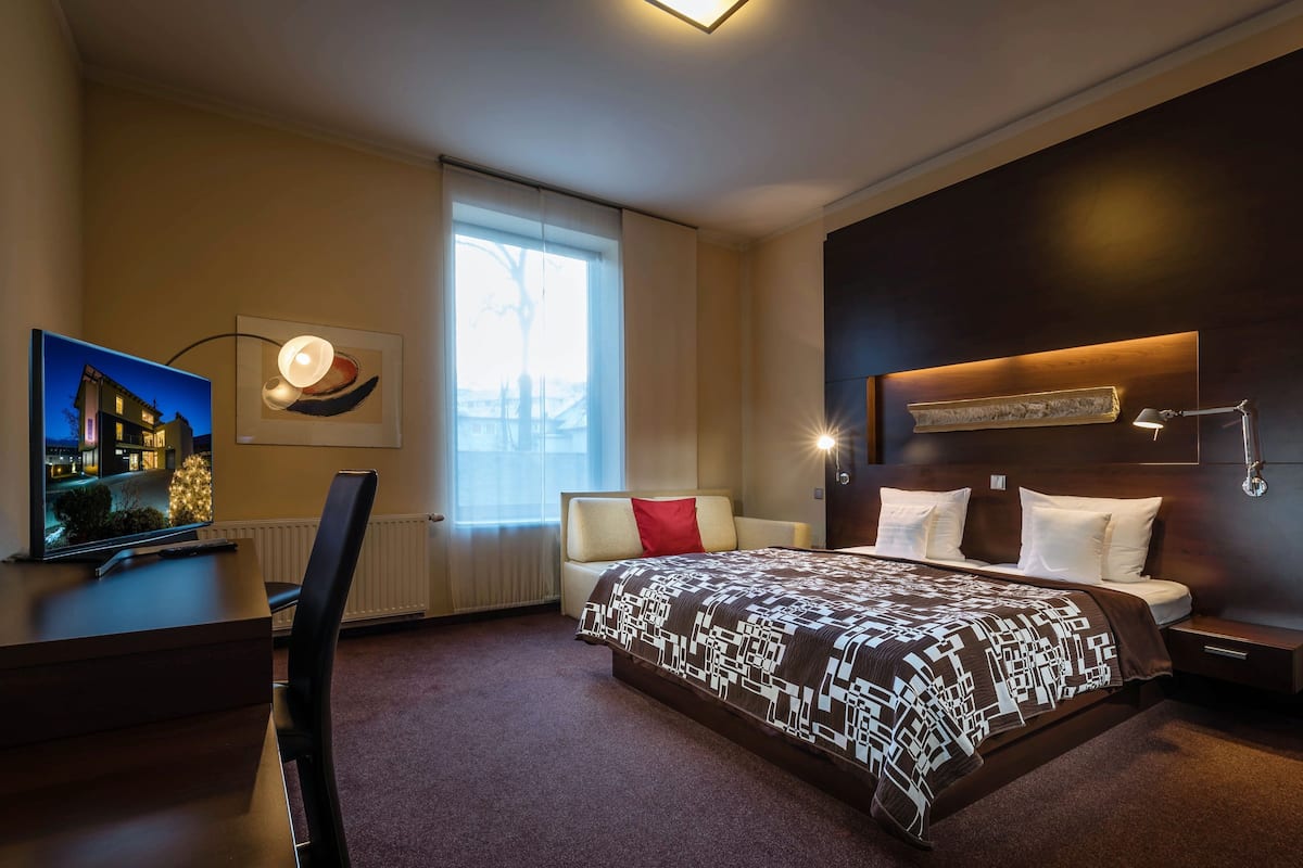 A modern hotel room with a large bed, a desk with a computer, a TV, and a window with curtains.