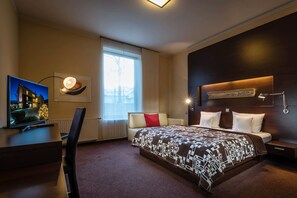 Una habitación de hotel moderna con una cama grande, un escritorio con computadora, televisor y ventana con cortinas.