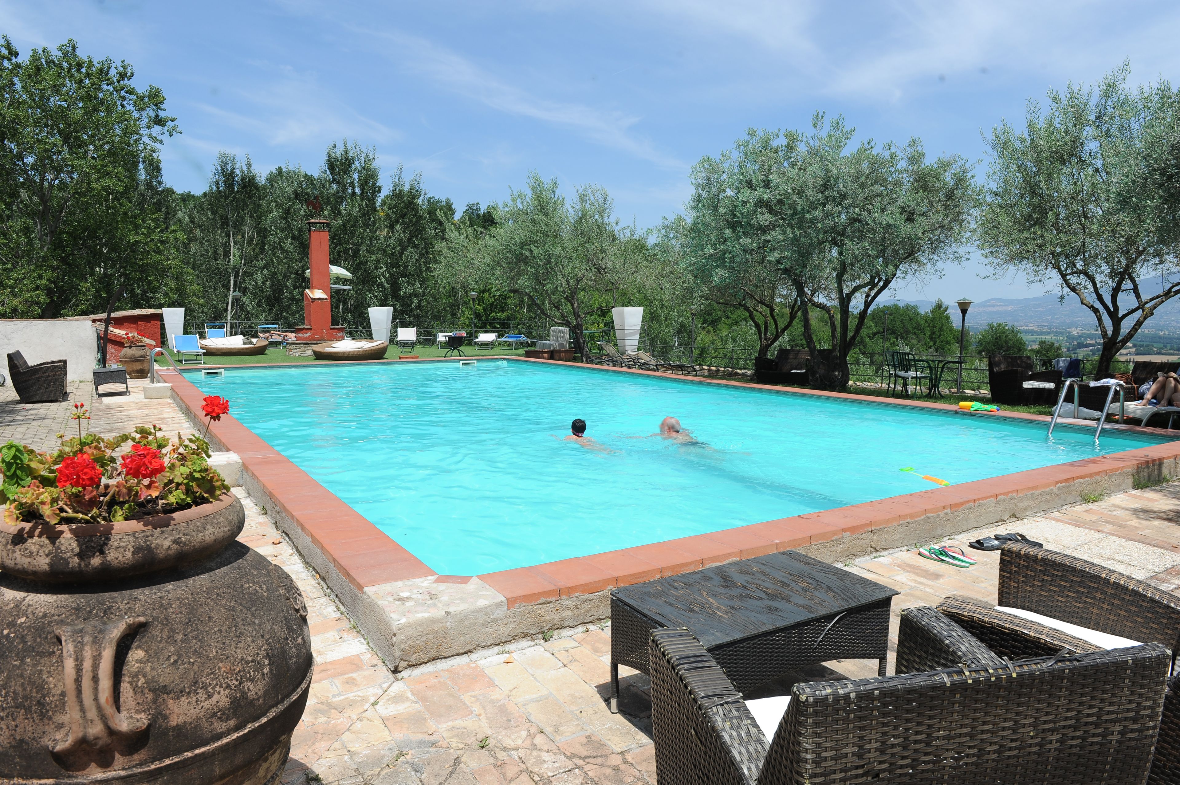 Una piscina con dos personas nadando, rodeada de mobiliario de exterior y plantas en macetas.