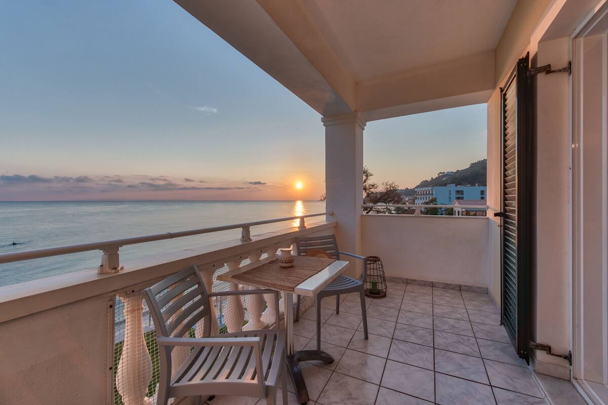 Un balcon avec vue sur la mer au coucher du soleil, comprenant une table et des chaises, une petite table avec une bougie et une lanterne décorative.