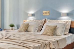 A neatly made bed with a beige blanket and patterned pillows, a small potted plant on a bedside table, and two wall-mounted light fixtures.