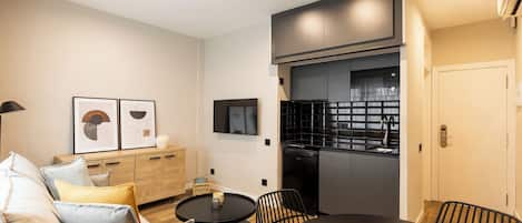 A modern living room with a sofa, a round coffee table, and a dining table with chairs. There is a kitchen area with a refrigerator and a microwave.