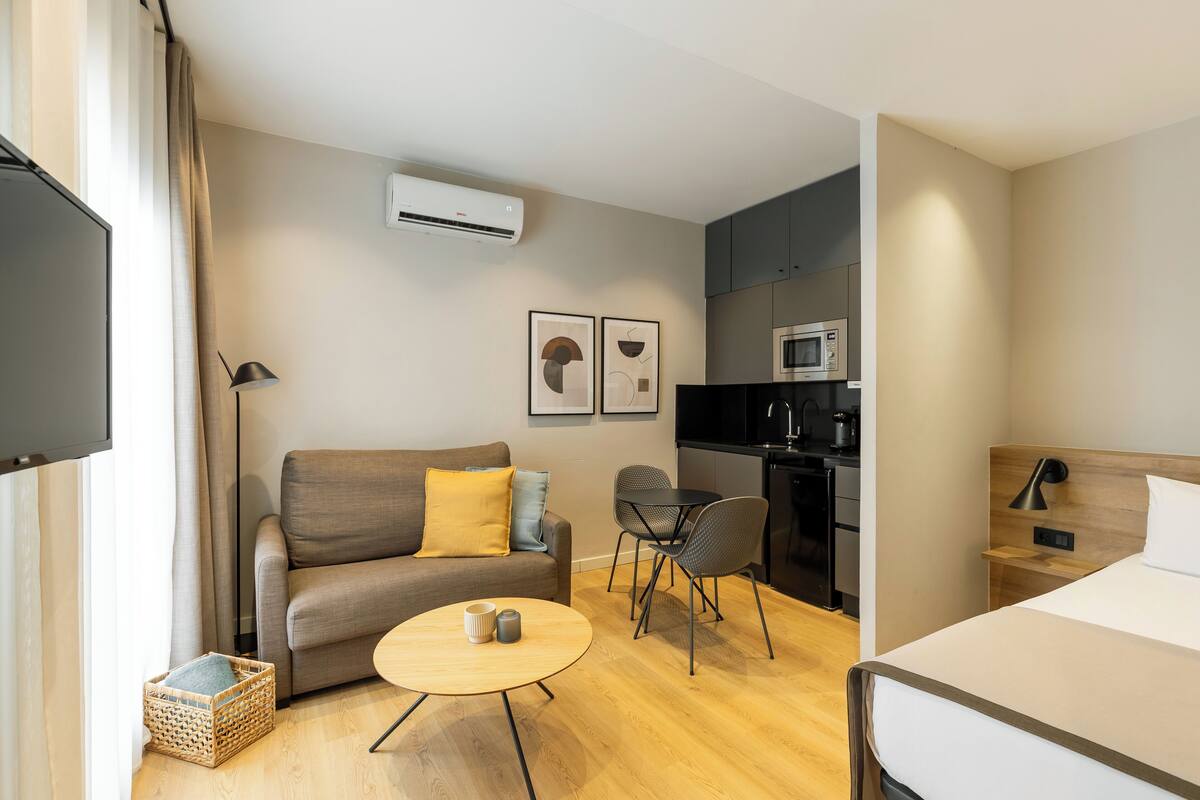 A modern hotel room with a sofa, a small round table, a bed, a kitchenette, and a TV.