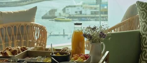 Una bandeja de desayuno con comida y bebidas sobre una mesa junto a una ventana con vista a un puerto deportivo.