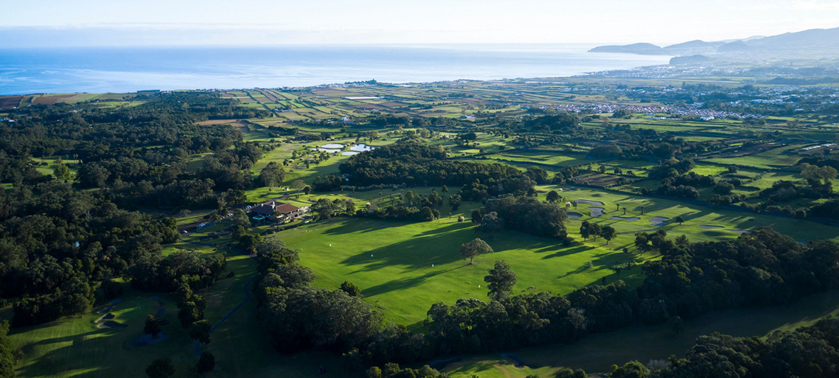 An aerial view of a golf course with a clubhouse, surrounded by lush greenery and a coastal backdrop.