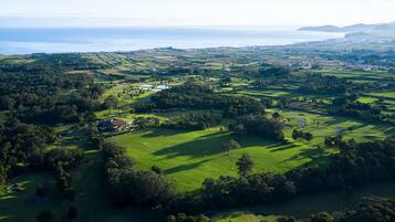 An aerial view of a golf course with a clubhouse, surrounded by lush greenery and a coastal backdrop.