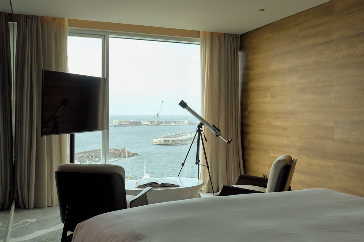 A hotel room with a large bed, a desk with a laptop, a chair, a TV, and a view of the water and a crane.