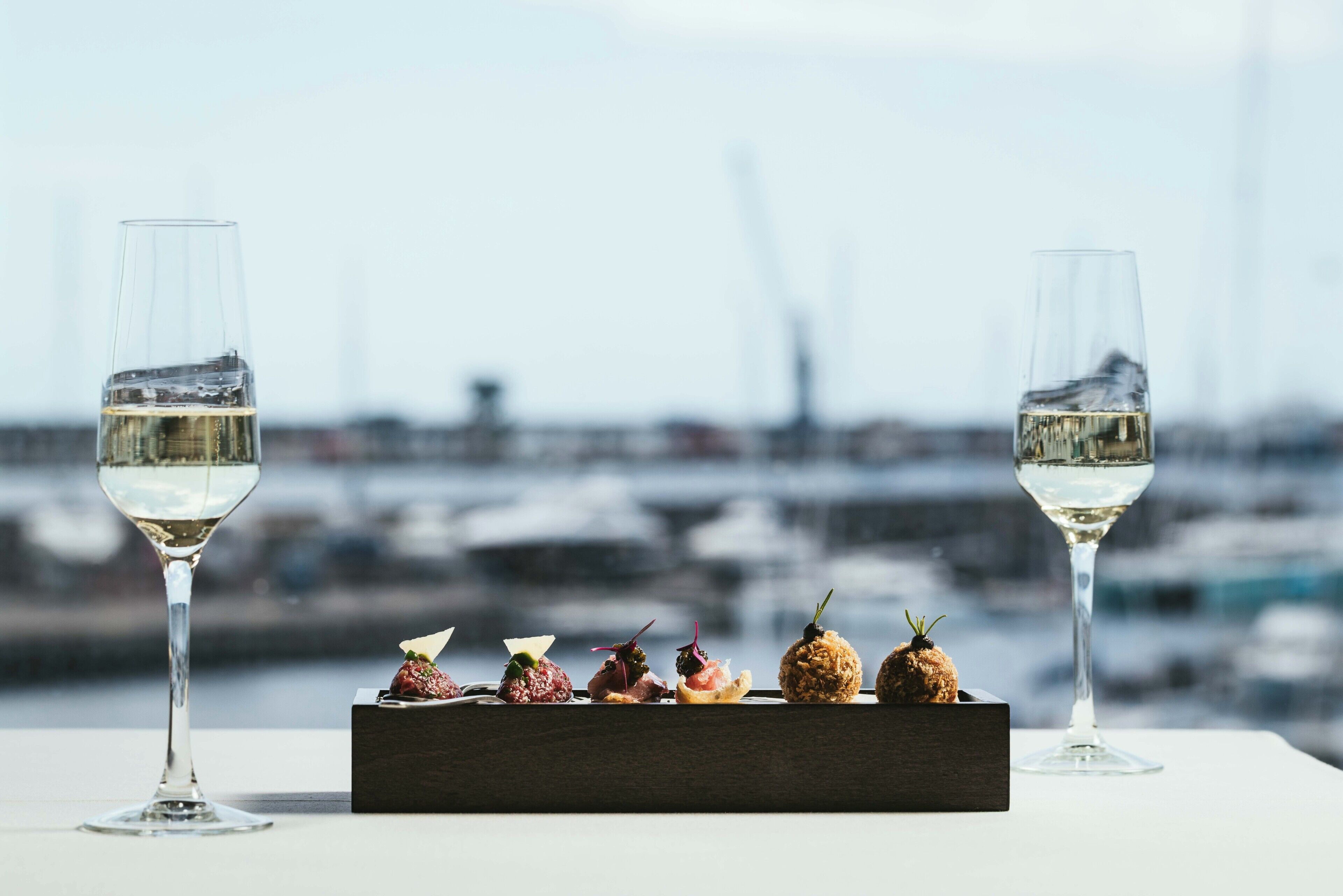 Una mesa con dos copas de vino y una caja de madera con diversos aperitivos.
