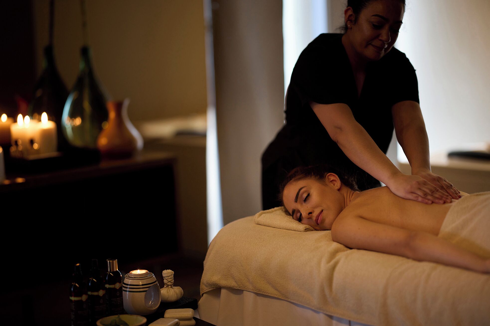 A massage therapy session in a dimly lit room with candles, bottles, and a towel-covered client.