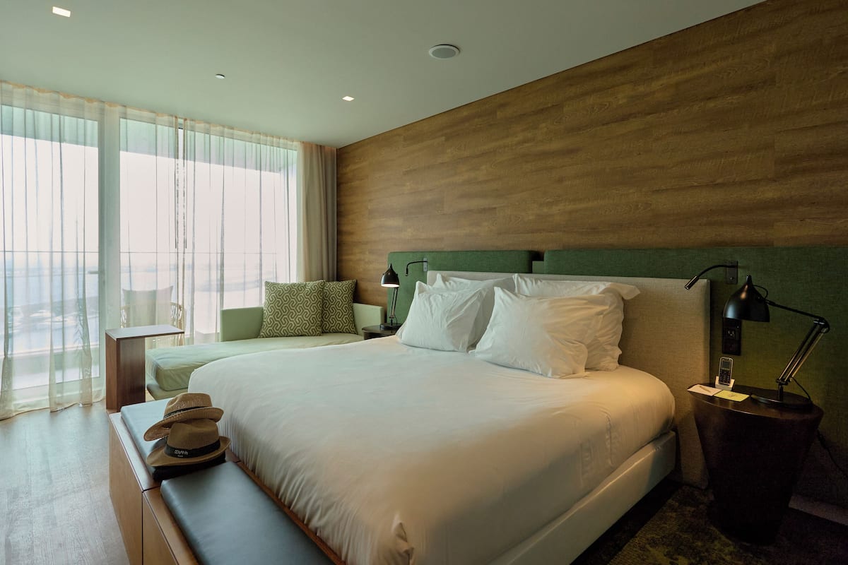 A hotel room with a large bed, a bedside table with a lamp, a small bedside table with a phone, a bench with a hat, and a window with sheer curtains.