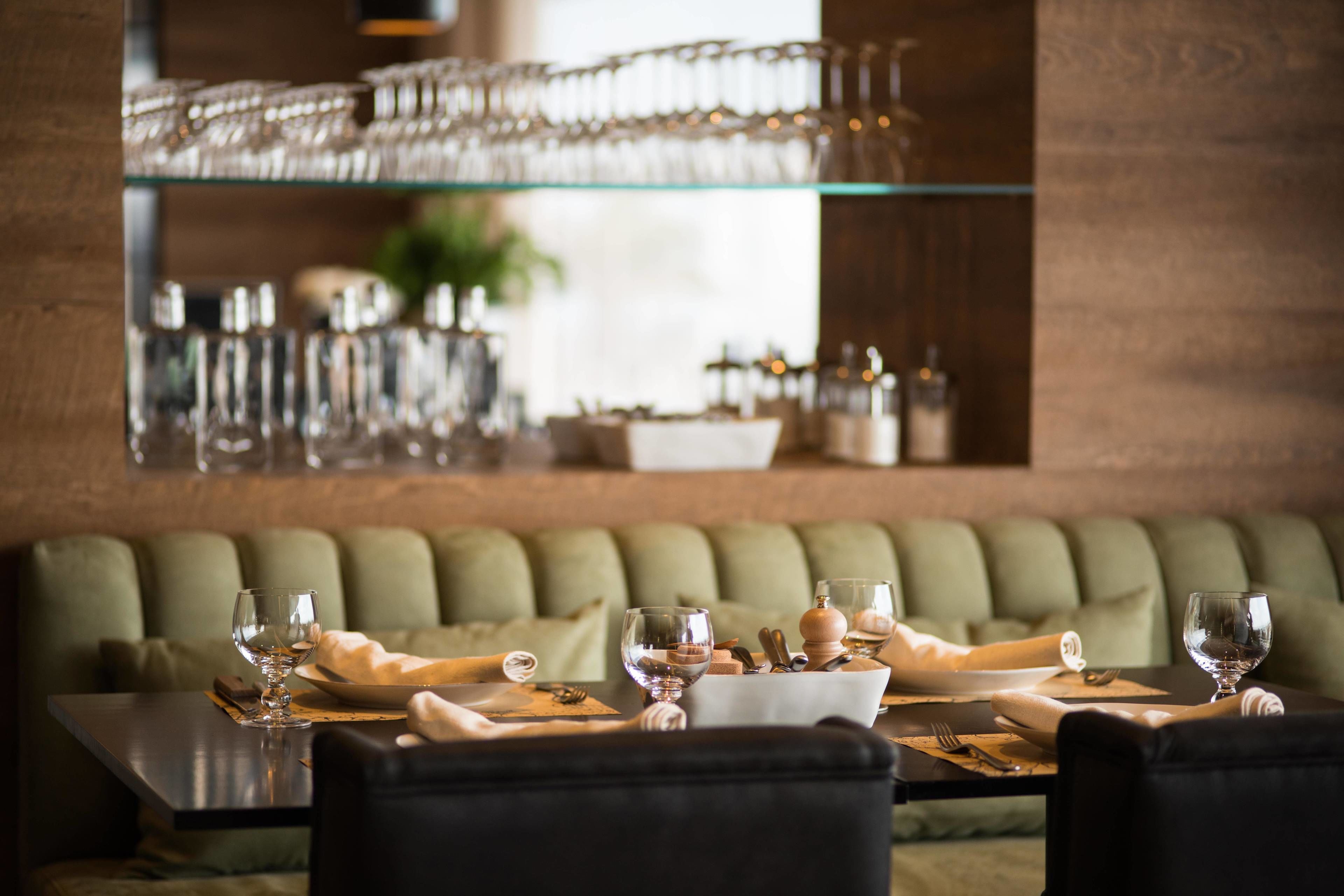 A dining area with a glass display of wine glasses on a shelf, a green cushioned bench, and a table set with plates, glasses, and cutlery.