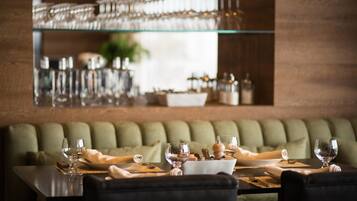 A dining area with a glass display of wine glasses on a shelf, a green cushioned bench, and a table set with plates, glasses, and cutlery.