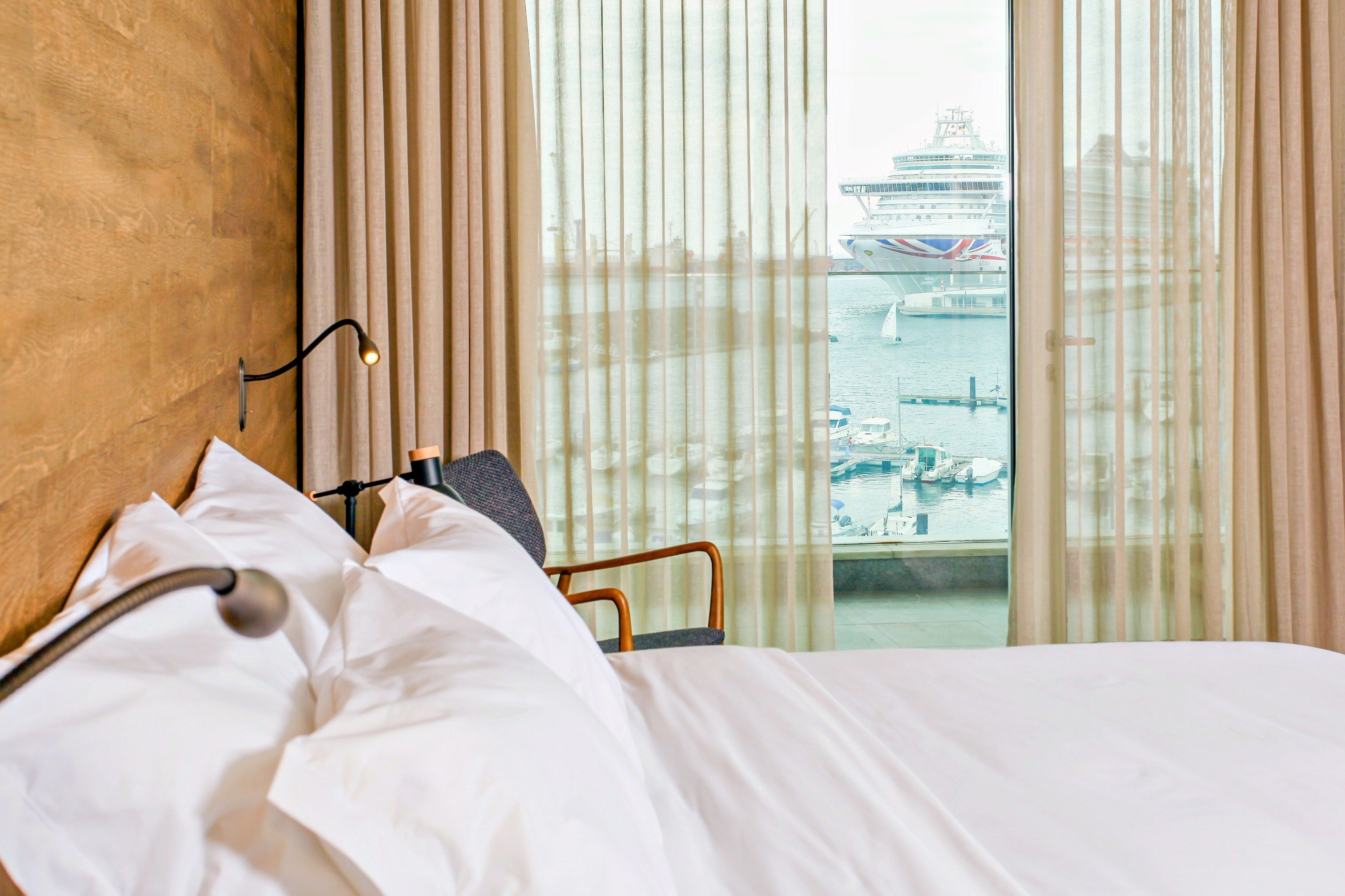 Habitación de hotel con cama, lámpara de noche y vista a un crucero y puerto deportivo.
