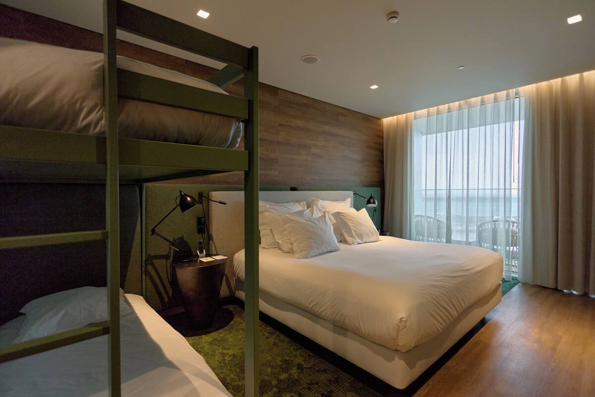 A hotel room with bunk beds, a large bed, a desk with a chair, and a window with curtains.