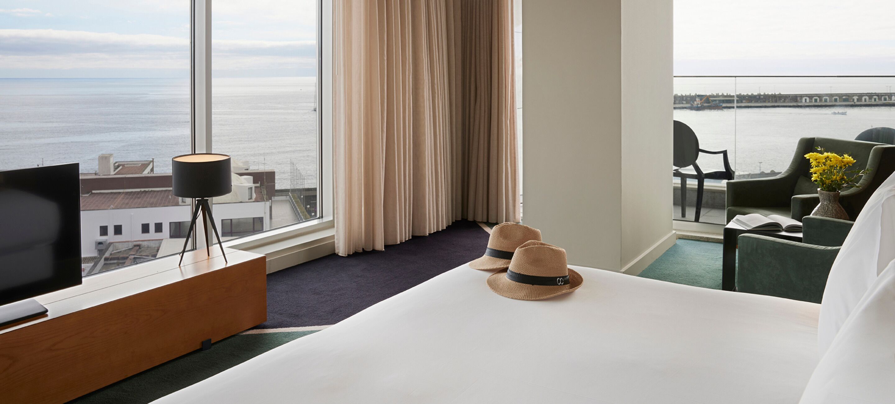 Habitación de hotel con un gran ventanal que ofrece vistas al mar y un puente, una cama con edredón blanco, un televisor sobre un mueble de madera, un escritorio con lámpara y dos sombreros sobre la cama.