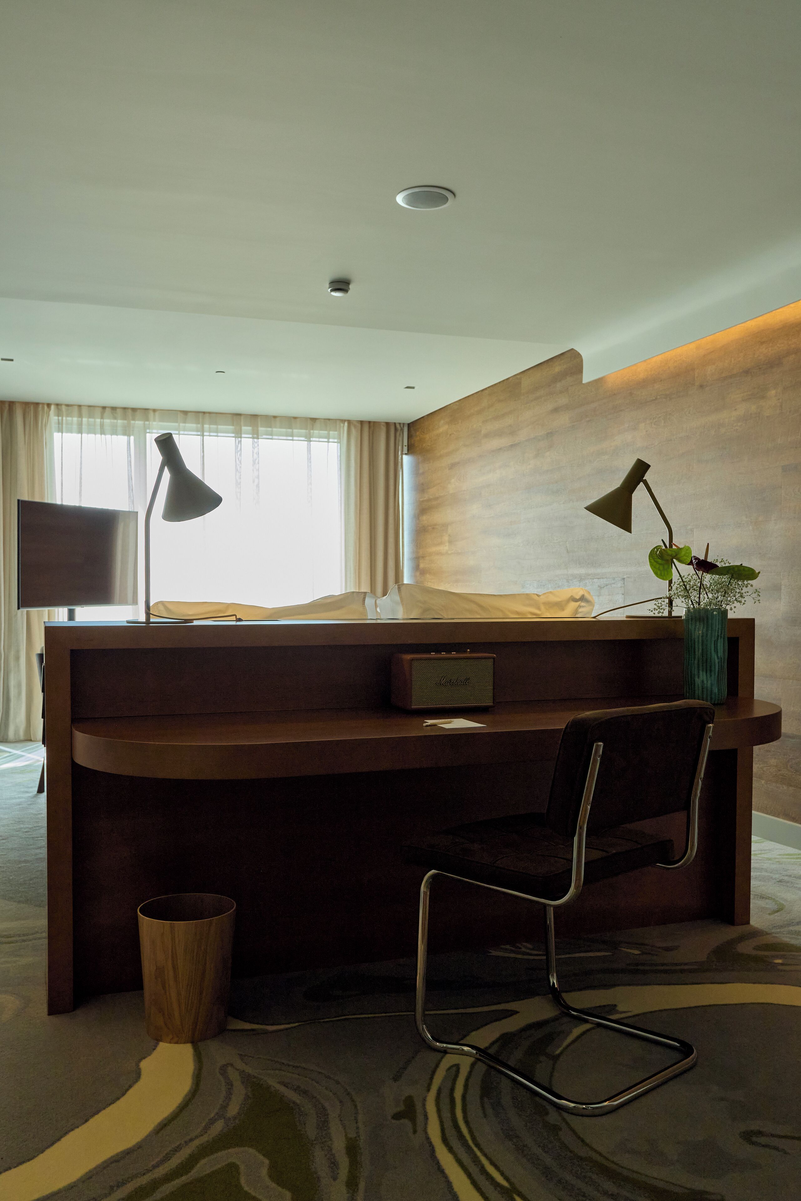 A modern hotel room with a wooden desk, two lamps, a chair, a TV, and a window with curtains.