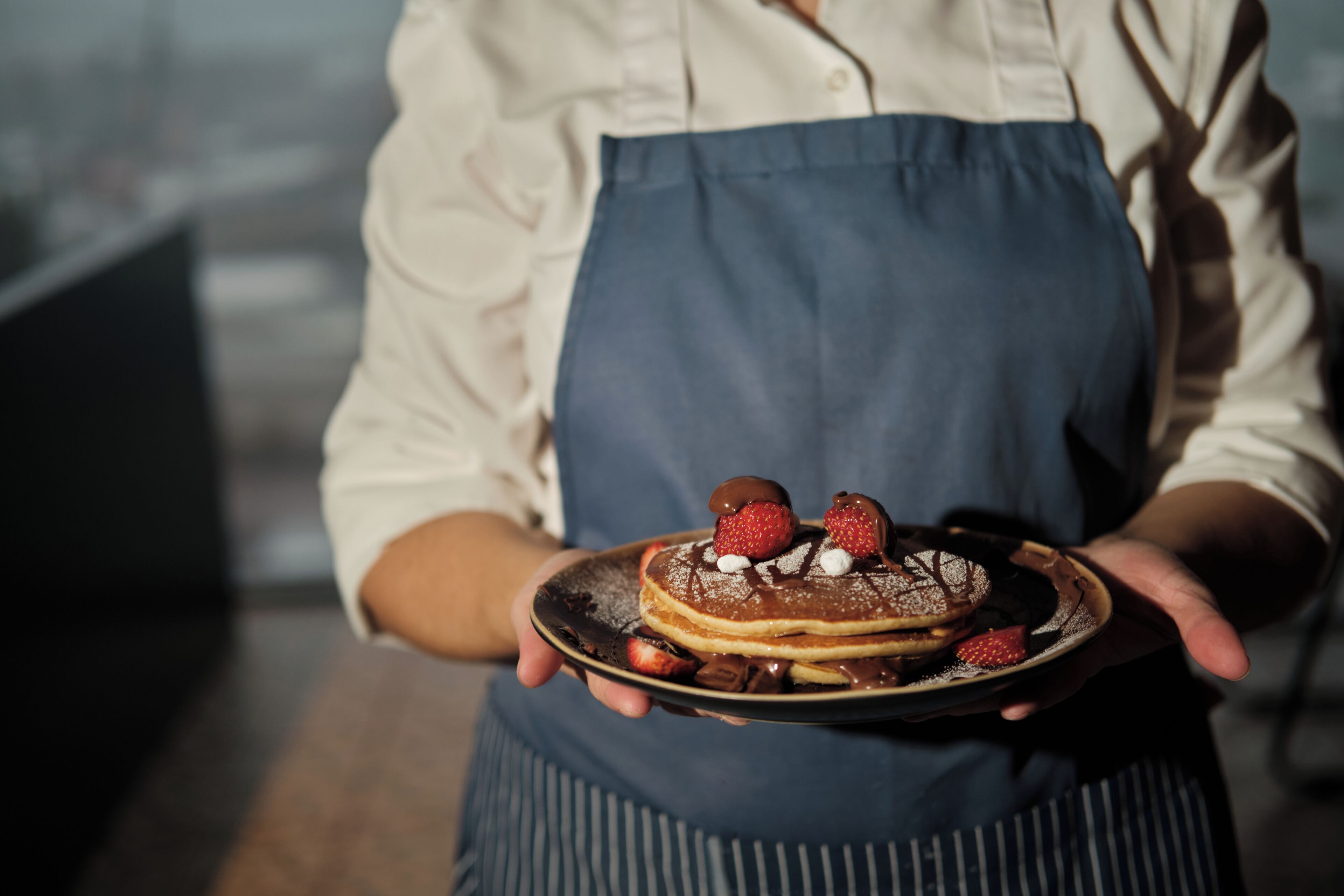 A person holding a plate with pancakes topped with strawberries and chocolate sauce.