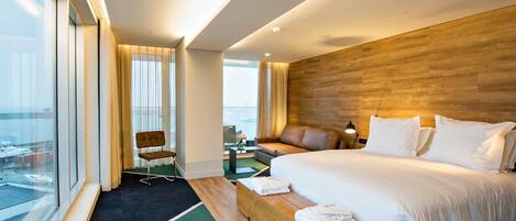 A modern hotel room with a large bed, a wooden headboard, a view of the city, and a seating area with a view.