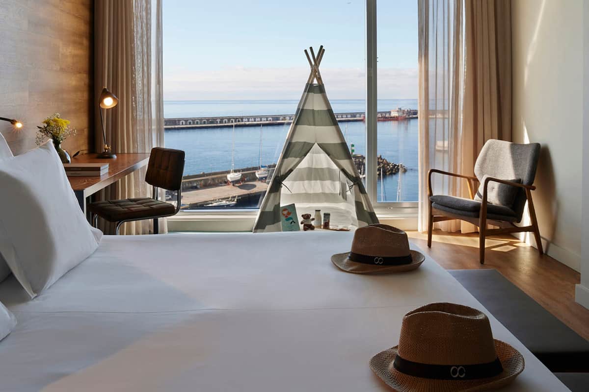 A hotel room with a large bed, a view of the sea, a wooden desk, a chair, and a teepee.