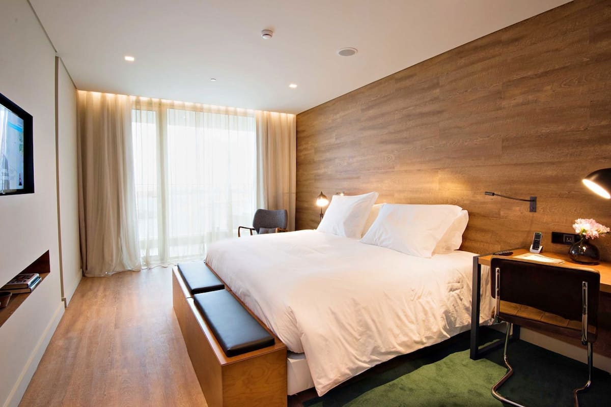A modern hotel room with a wooden accent wall, a large bed with white linens, a bench, a desk with a lamp, and a TV on the left wall.