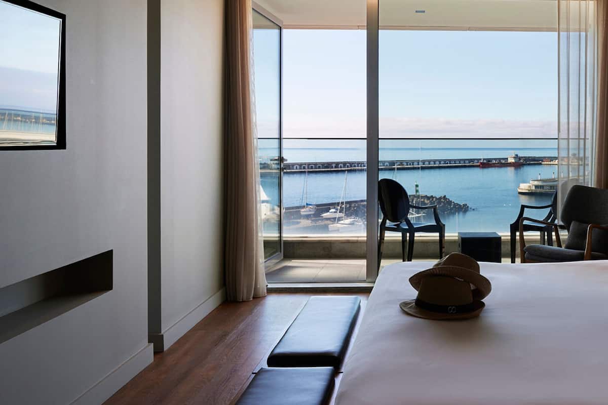 A hotel room with a large bed, a view of the sea and a pier, a small table with chairs, and a lamp.