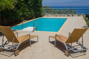 A rectangular swimming pool with a metal ladder, two wicker lounge chairs, and a stone wall with a metal railing.