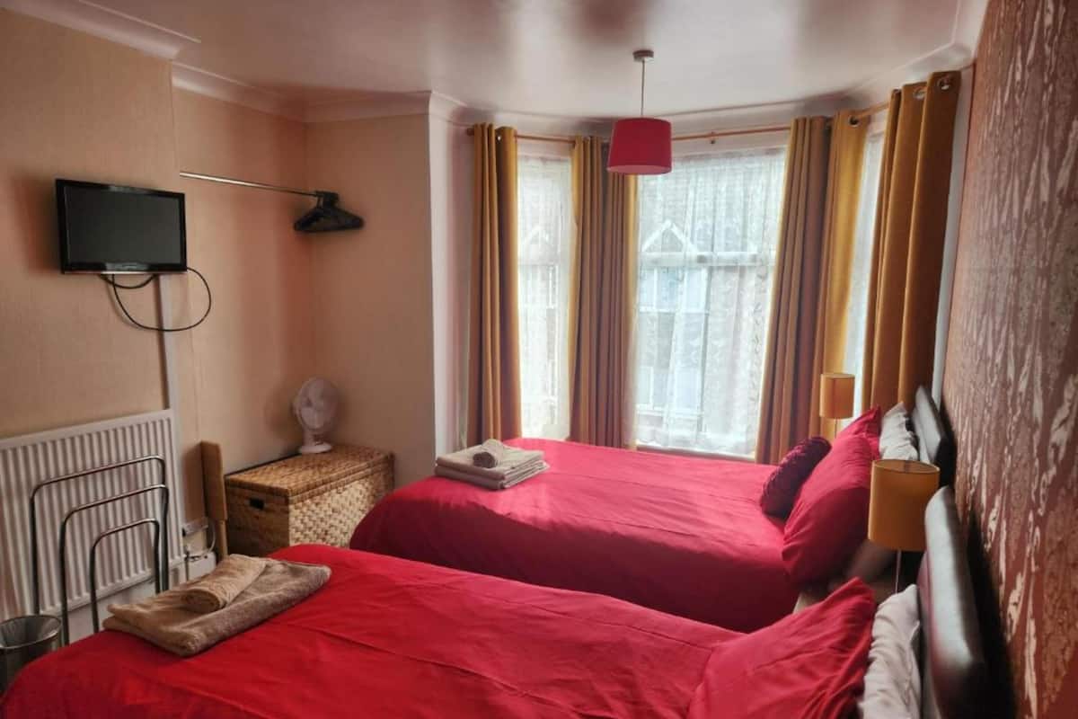 A hotel room with two beds, a television mounted on the wall, a radiator, and a window with curtains.