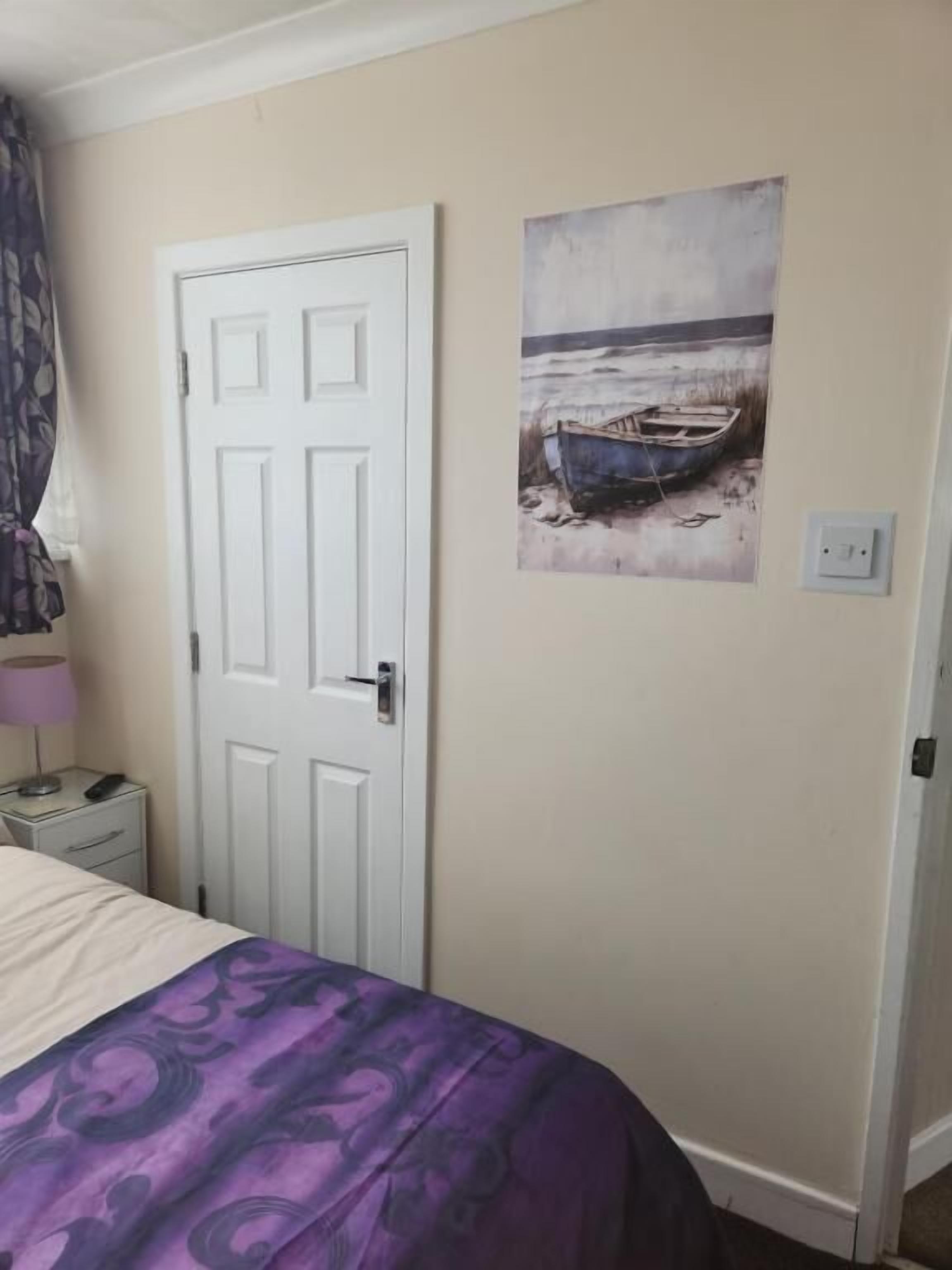 Um quarto com uma cama, uma porta, uma mesa de cabeceira, uma luminária e uma pintura de um barco na parede.
