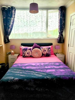 Un dormitorio con cama, dos mesitas de noche, una ventana con cortinas y una lámpara colgante.