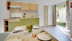 A compact kitchen with wooden cabinets, a dining table set for four, and a view of the outdoors through a sliding door. - Camping Fusina (Mestre)