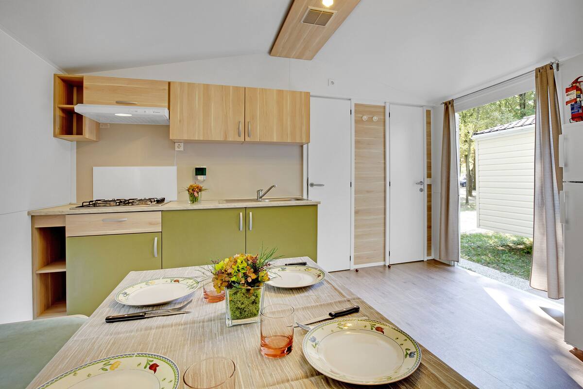 A compact kitchen with wooden cabinets, a dining table set for four, and a view of the outdoors through a sliding door.