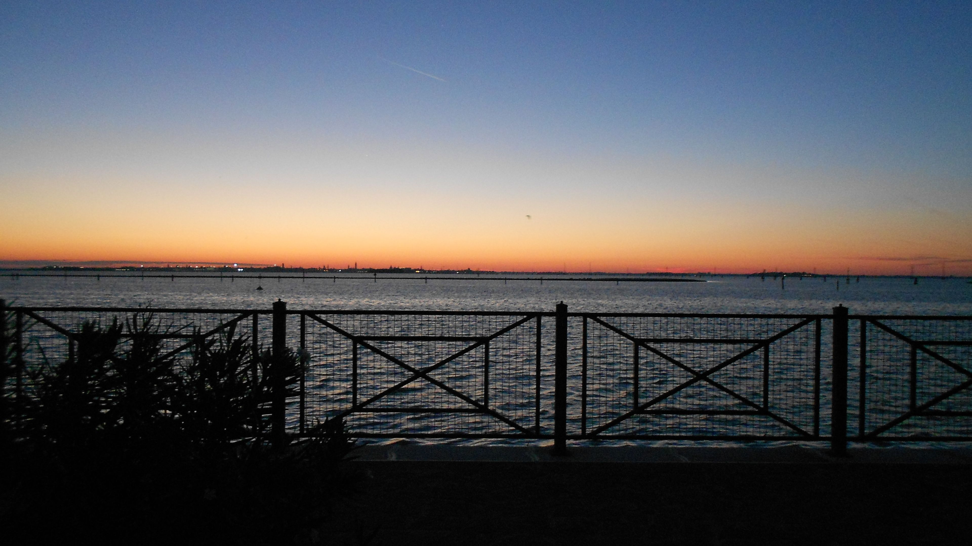 Une vue sur le bord de l’eau, avec une clôture en métal, une eau calme et un ciel au coucher du soleil.