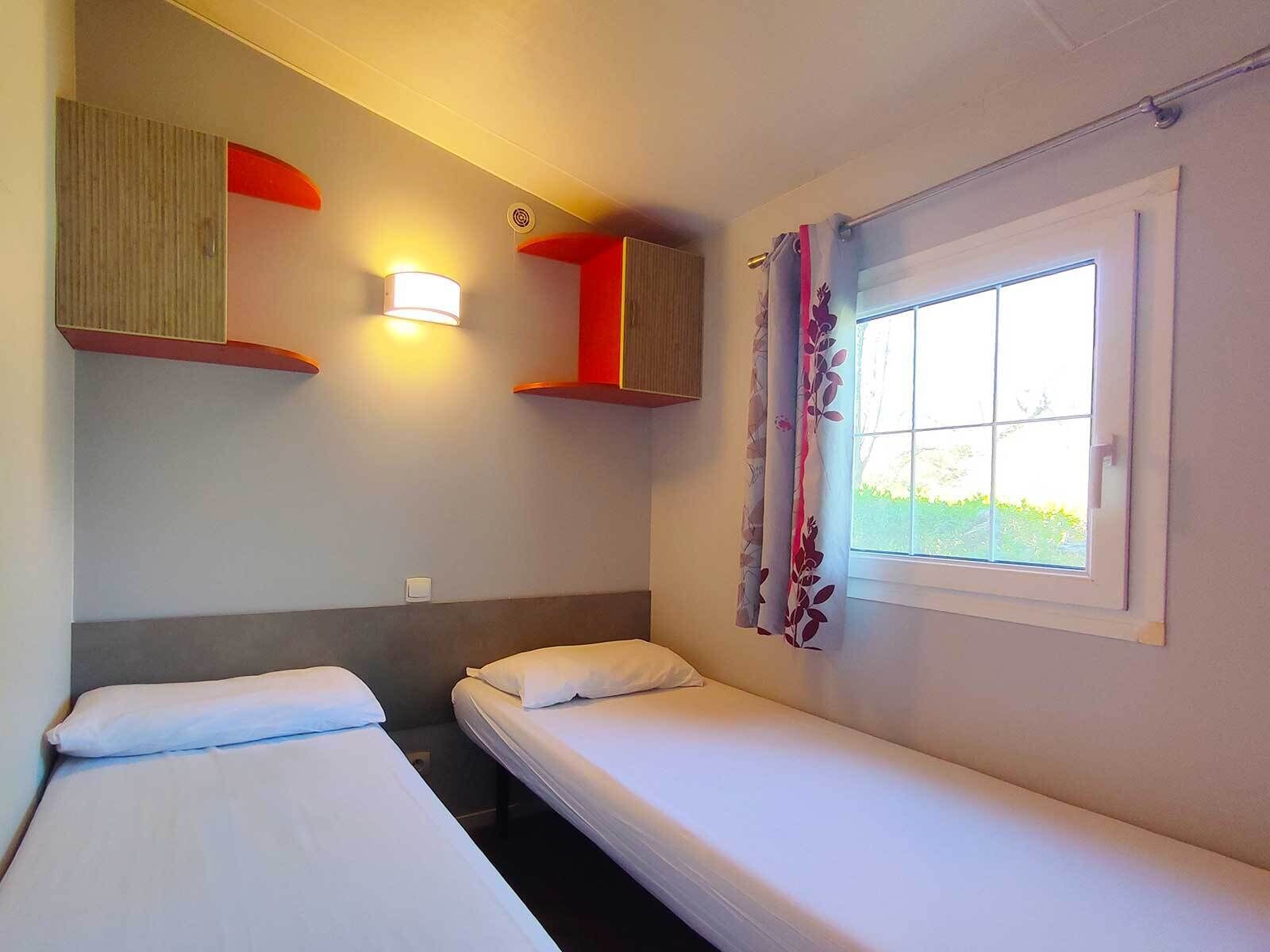 A small, simple room with two single beds, a window with curtains, and a wall-mounted light fixture.