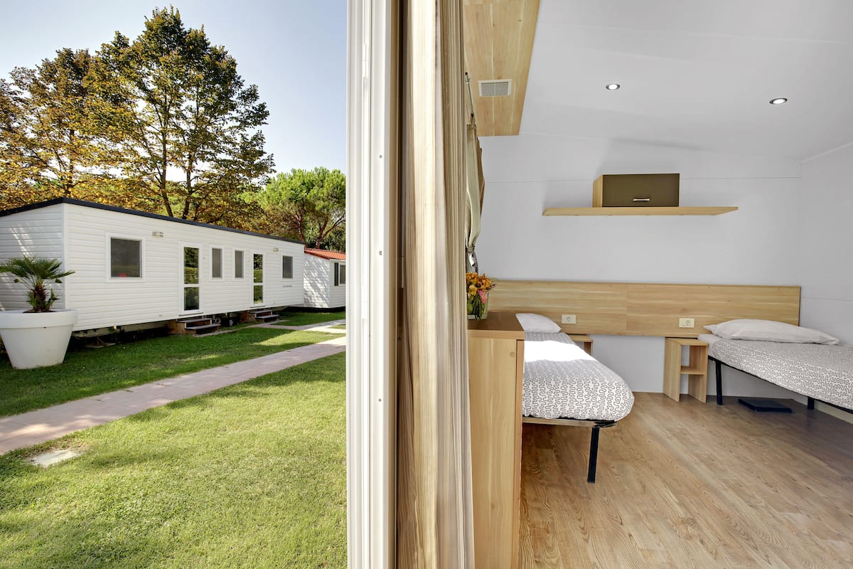 A modern, well-lit room with two beds, a small desk, and a view of a white mobile home in the background.