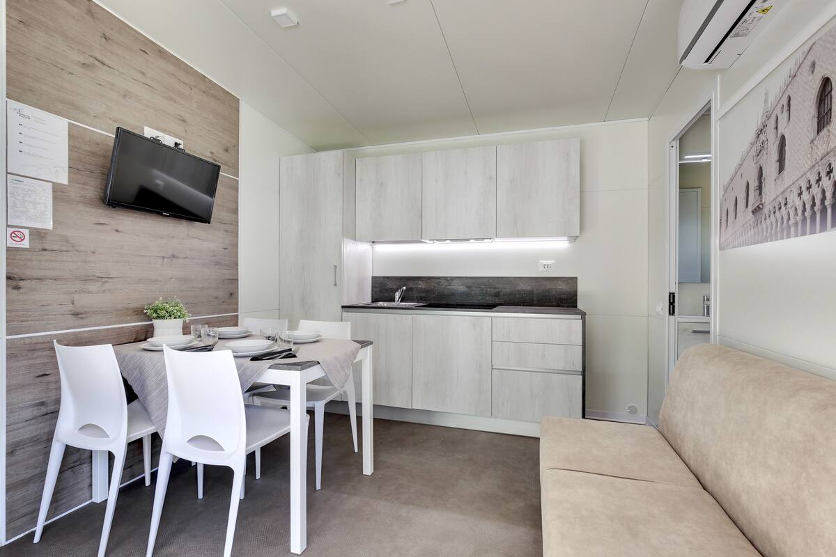A compact kitchen with a built-in stove and white cabinetry, a dining area with a table and chairs, and a beige sofa.