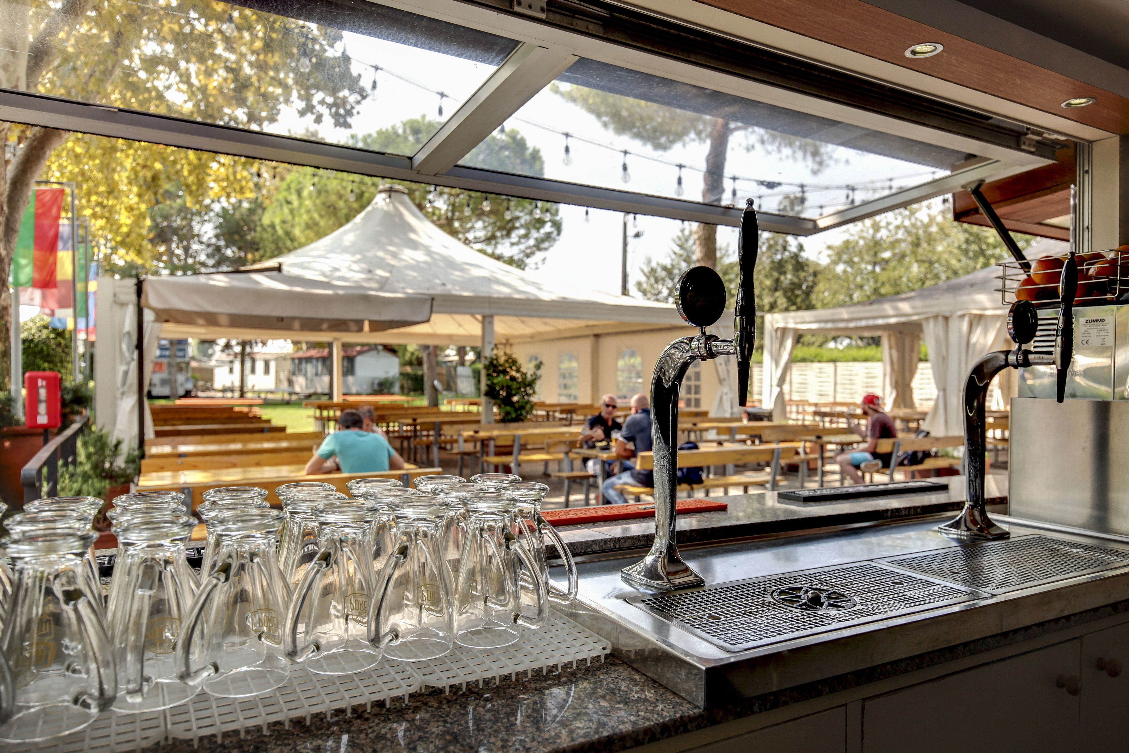 A bar area with a stainless steel sink, a glass mug rack, and a view of an outdoor seating area with tables and umbrellas.