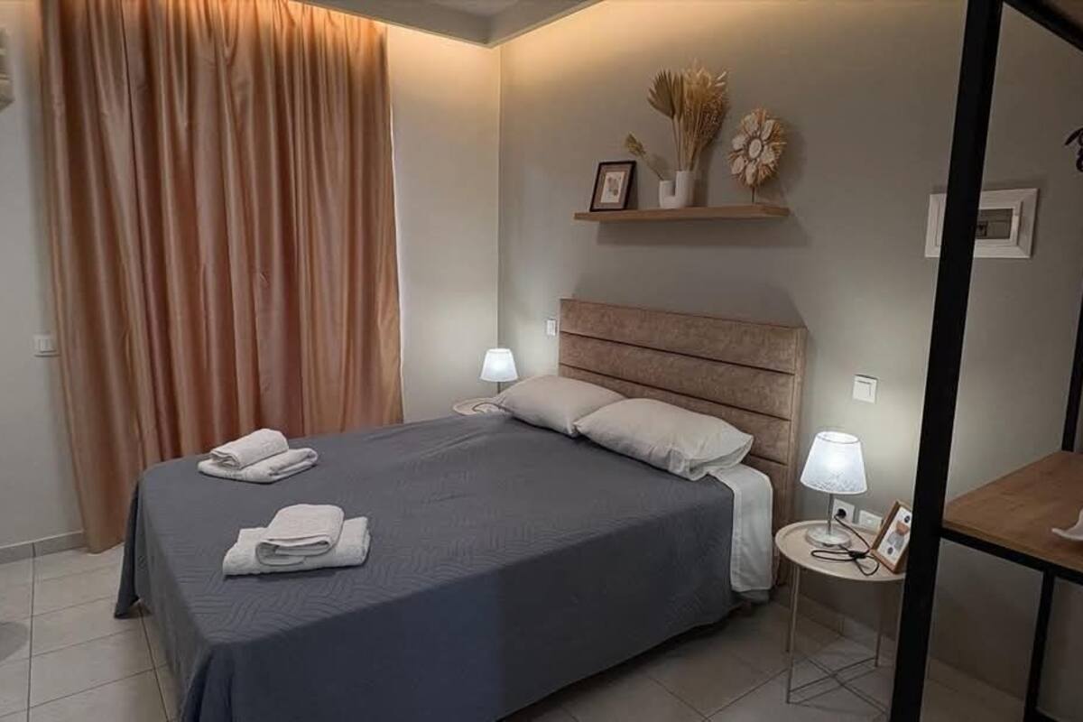 Una cama bien hecha con colcha azul oscuro y almohadas blancas, una mesita de noche con lámpara, y un pequeño estante con objetos decorativos.