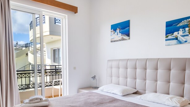 Una cama hecha con pulcritud, con un edredón marrón, almohadas blancas y toallas dobladas. Hay dos cuadros en la pared que representan escenas costeras.