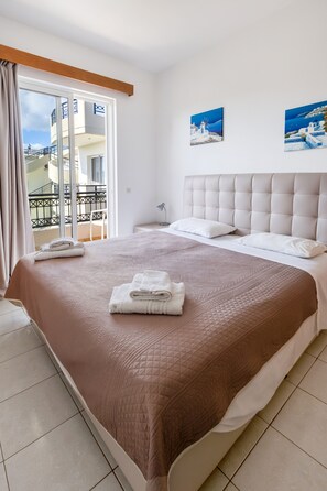 A neatly made bed with a brown quilt, white pillows, and folded towels. There are two paintings on the wall depicting coastal scenes. - Johnhara Studios & Apartments (Rhodes)