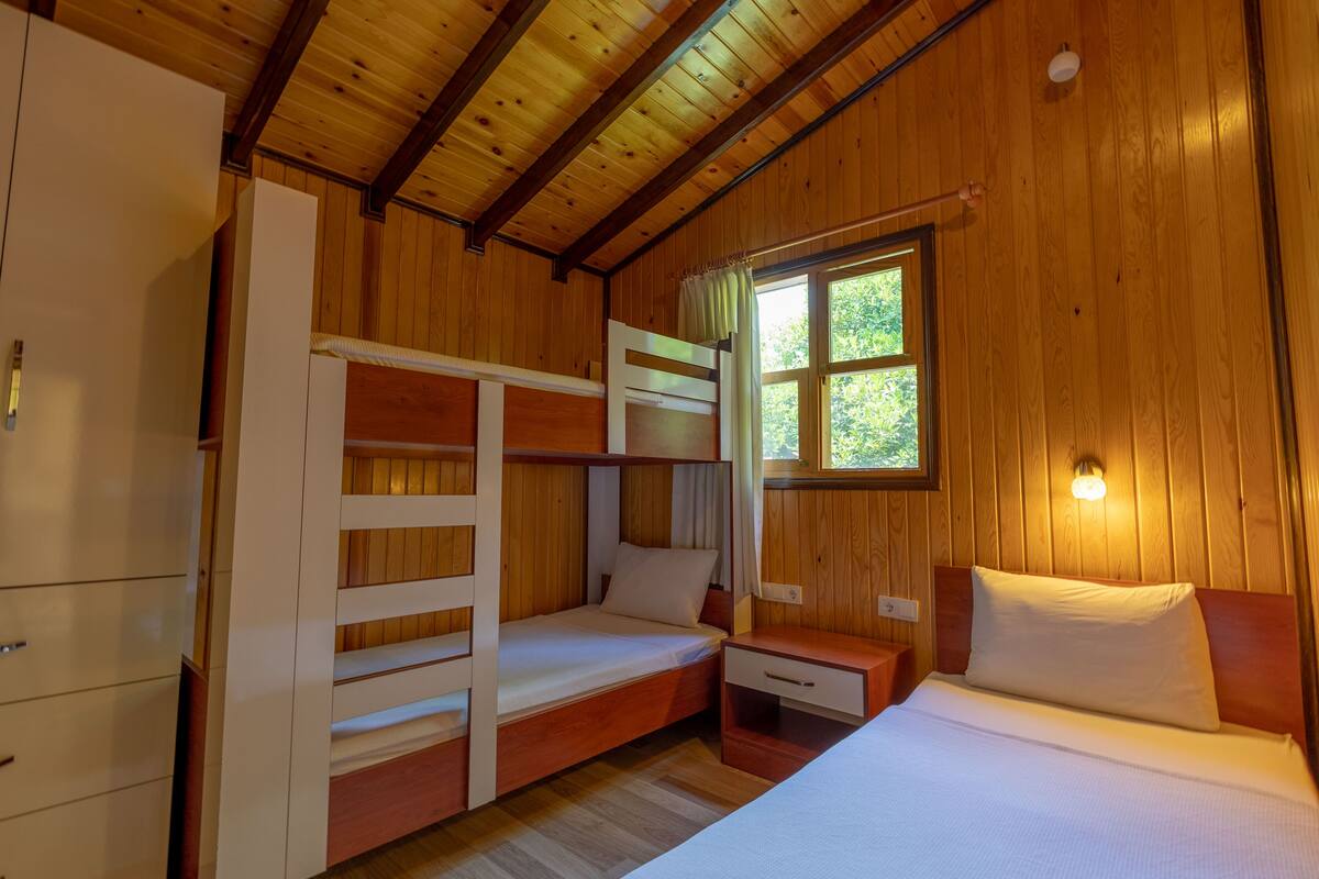 A wooden cabin room with bunk beds, a single bed, a nightstand, and a window with a view of trees.