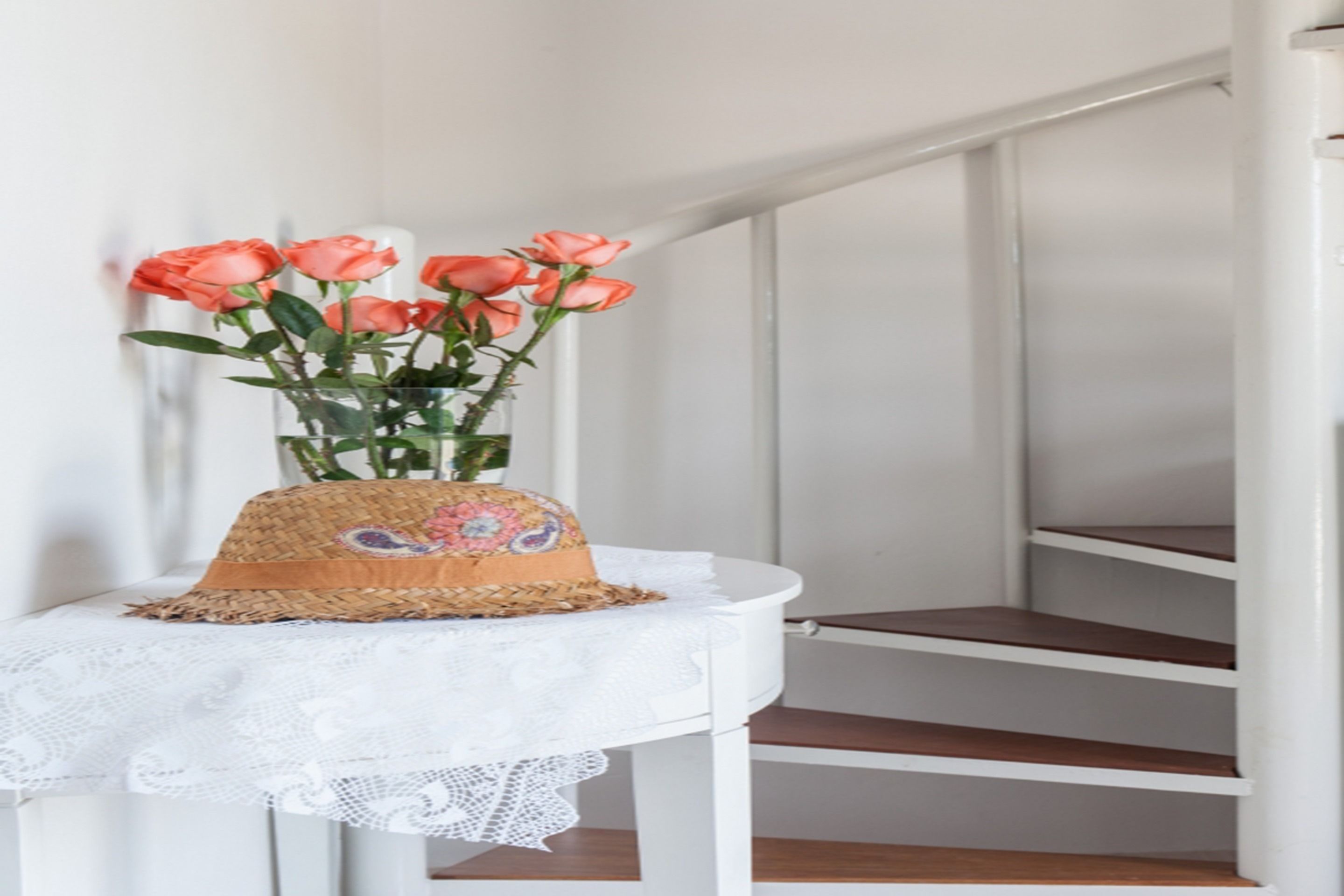 Una escalera con barandilla blanca y una mesita con un sombrero tejido y un florero de rosas rosadas.