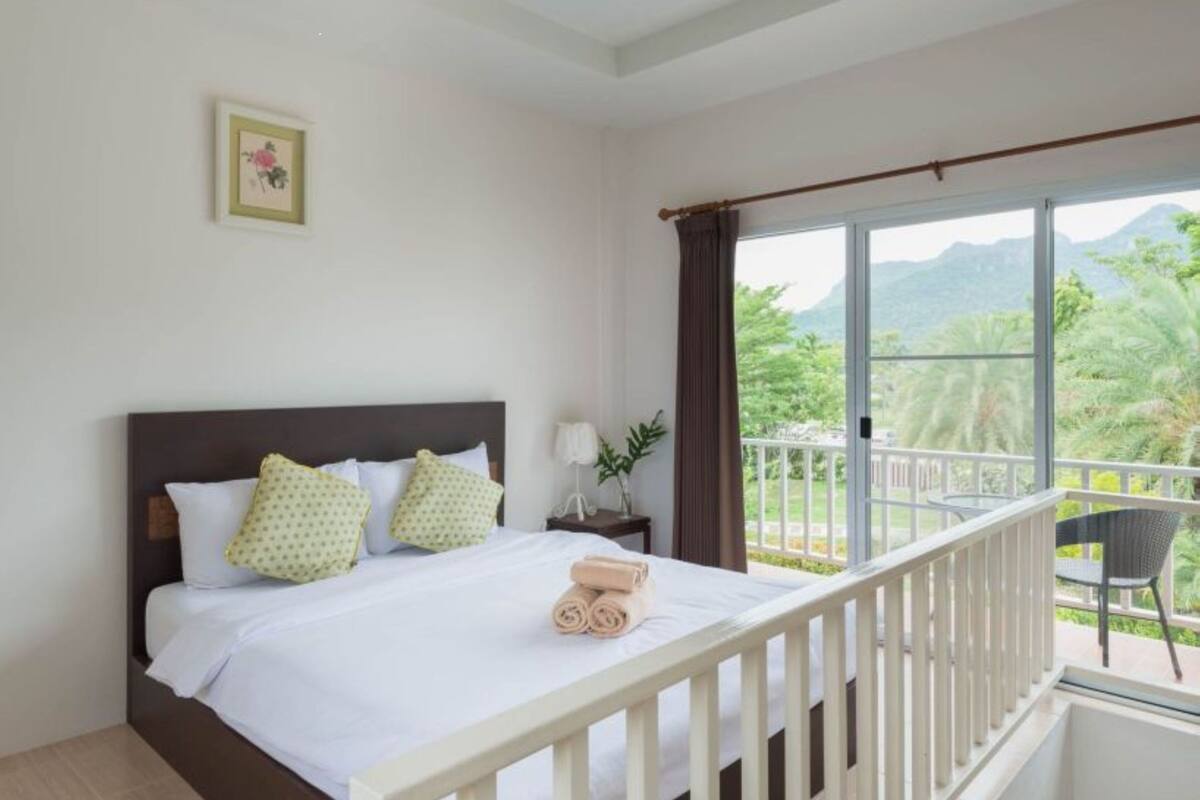 Ein Hotelzimmer mit Bett, Nachttisch, Fenster mit Blick auf Grünflächen und Balkon mit einem Stuhl.