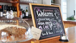 A chalkboard sign with a welcome message, a cake display, and a selection of drinks including gin and tonic. - Rosengaarden Hostel (Akirkeby)