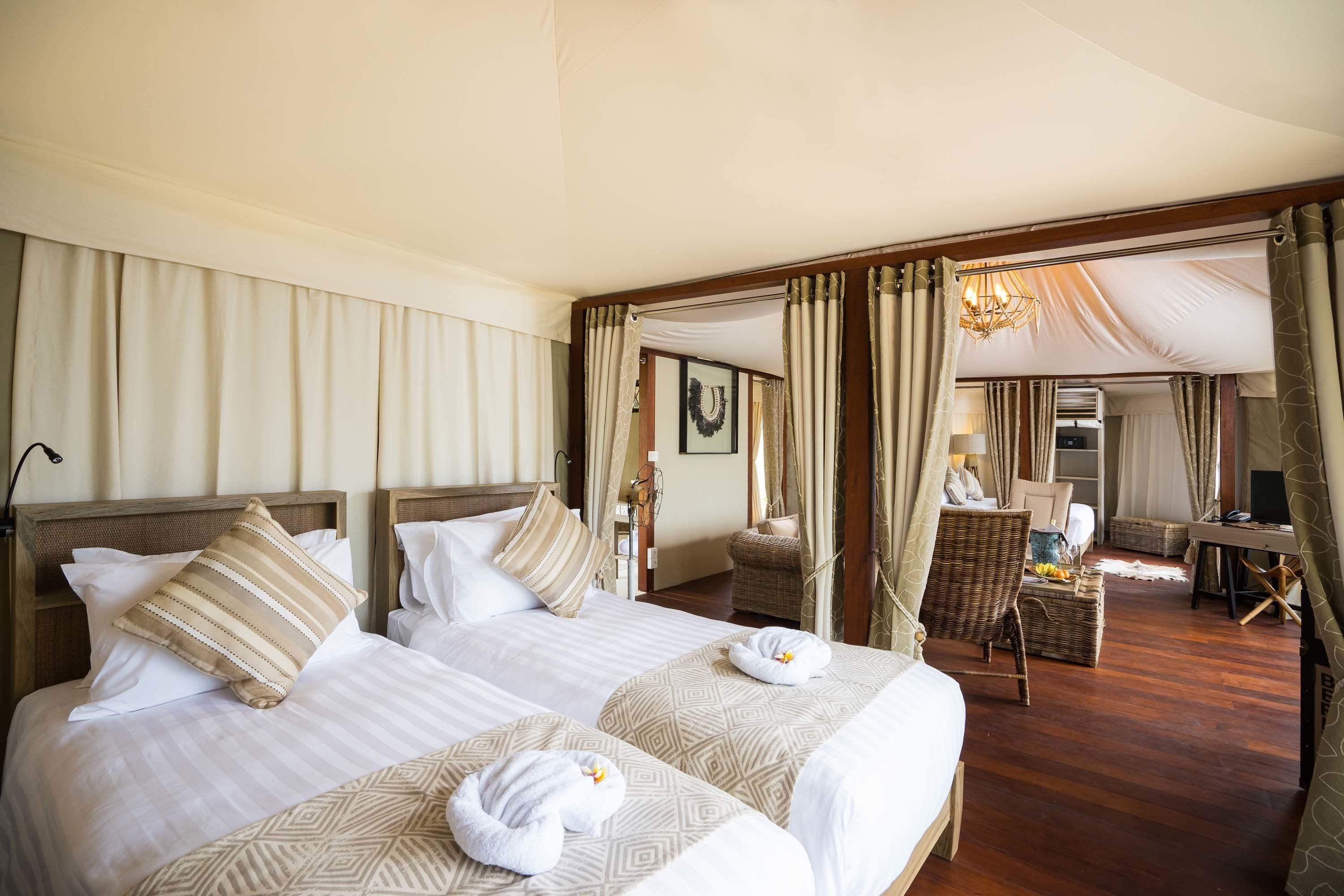 Quarto de hotel com duas camas, um espaço de estar com uma poltrona e mesa, e vista para o exterior.