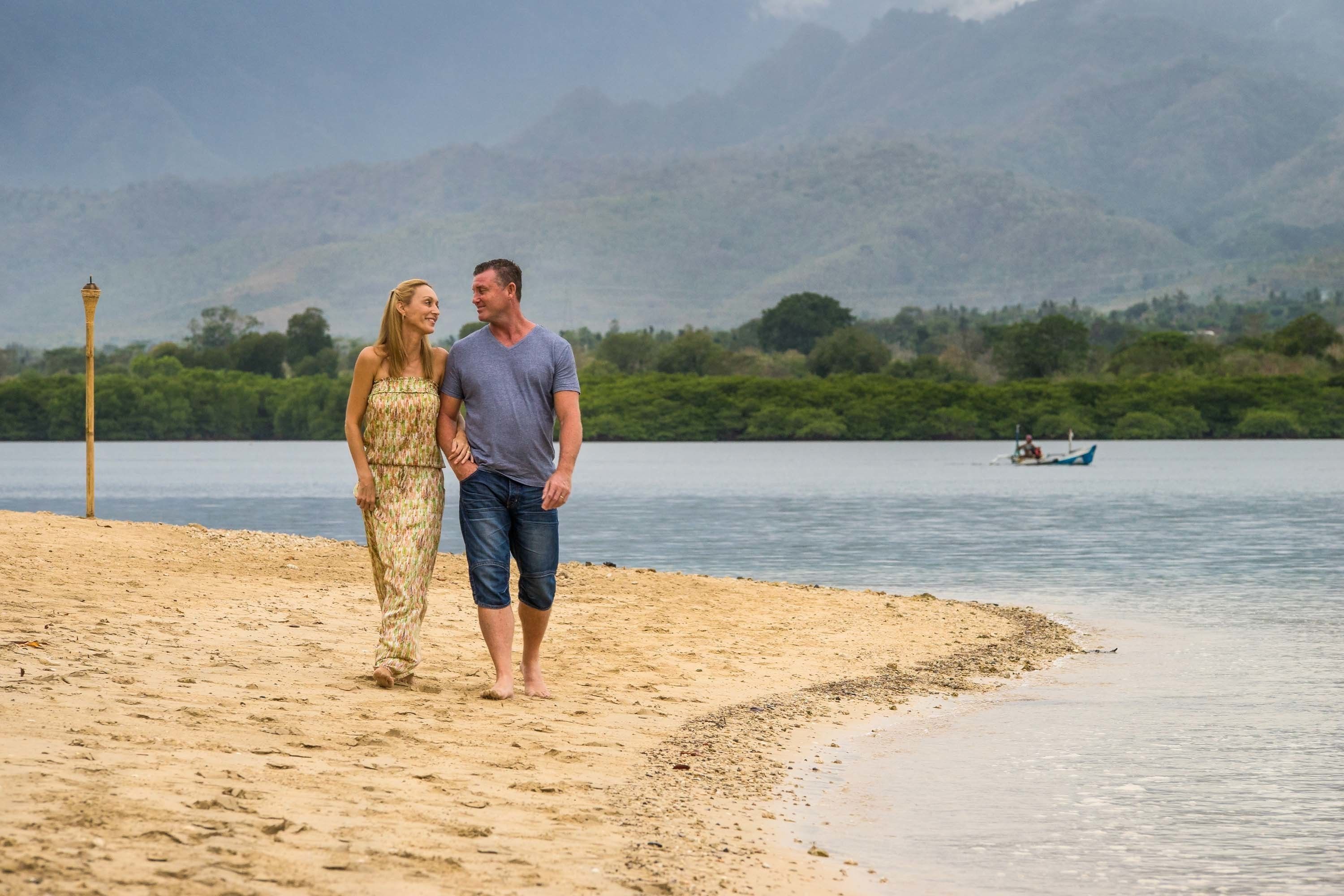 Una pareja caminando sobre una playa arenosa con un barco al fondo y montañas a lo lejos.