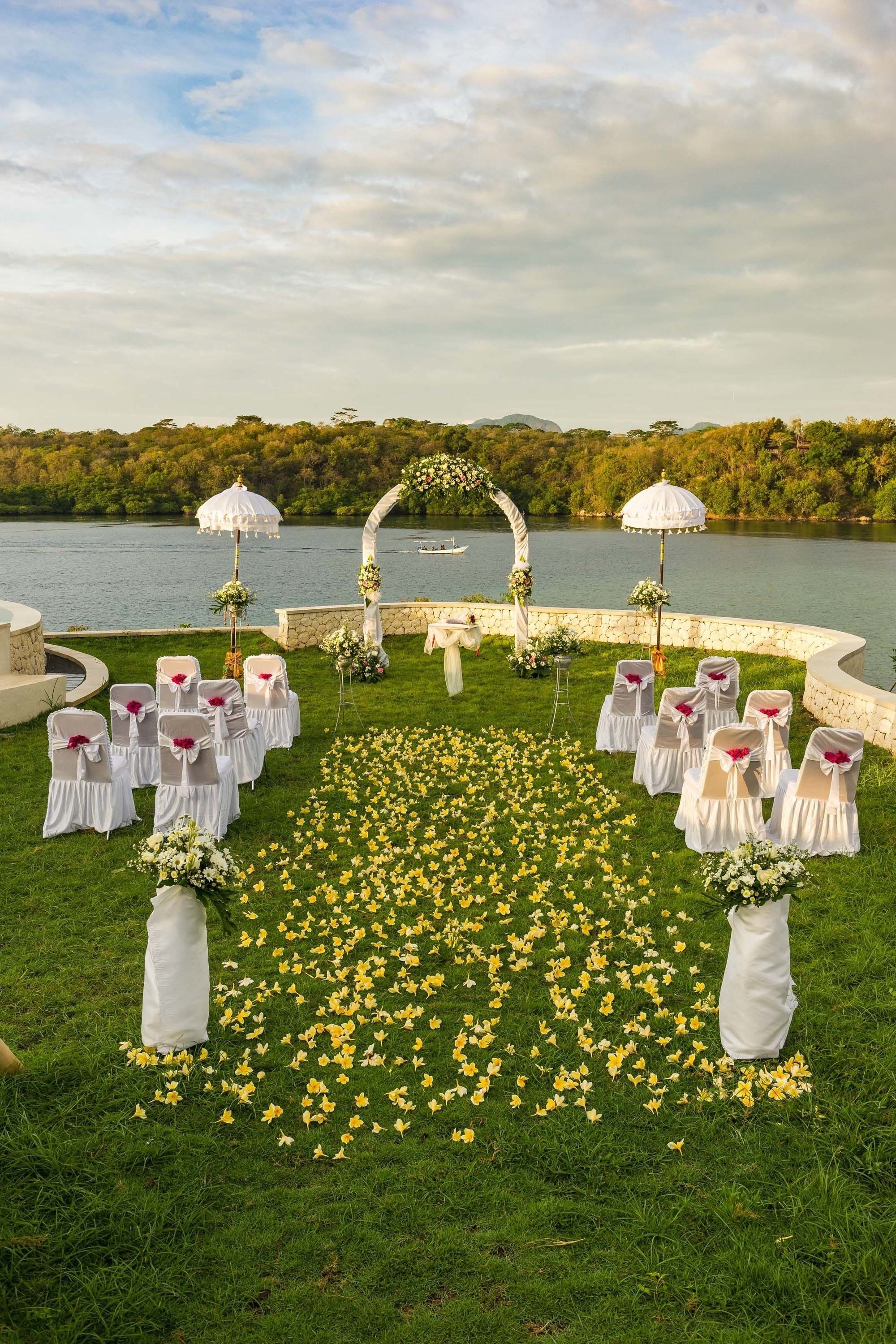An outdoor wedding setup with a lake view, white chairs with red sashes, and a floral arch.